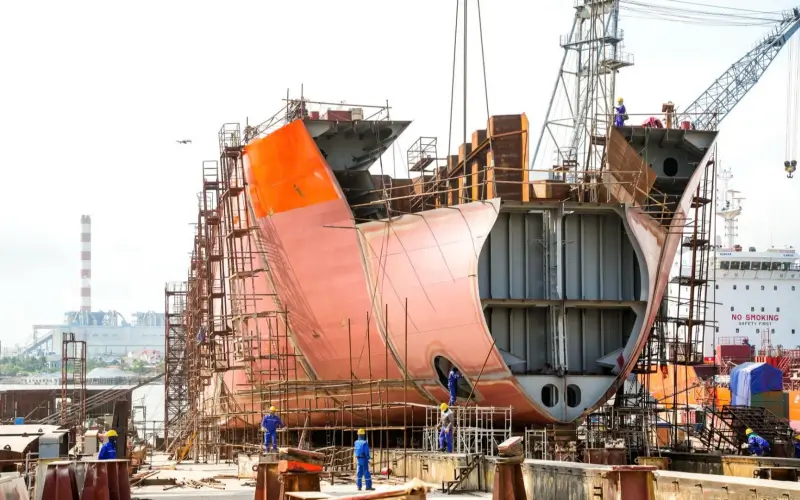 A massive cargo ship under construction in a shipyard with steel scaffolding and cranes, representing stud welding applications in maritime manufacturing.