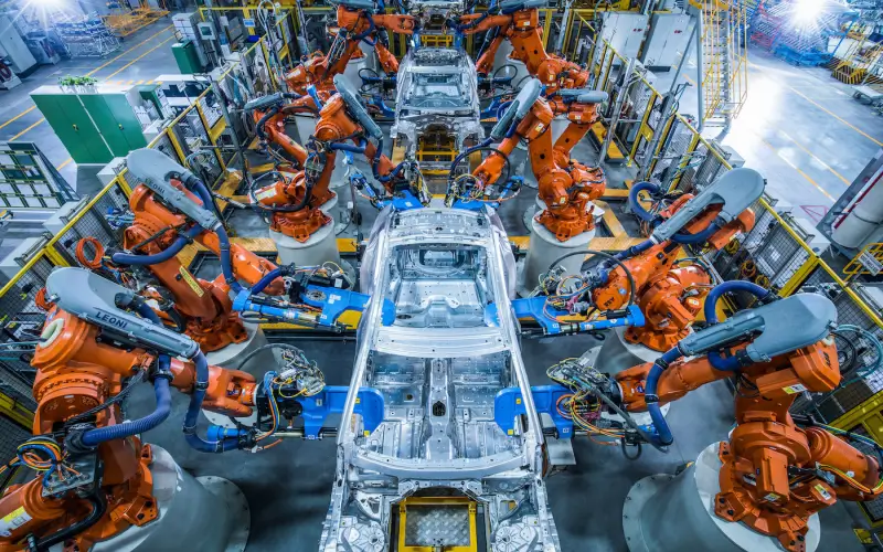 A modern automotive assembly line featuring orange robotic arms performing precision welding on a car chassis.